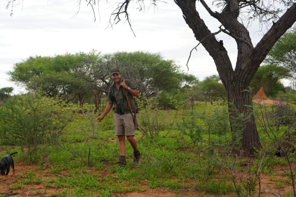 Jäger mit Flinte und Hund lächelnd im grünen Busch von Namibia – nachhaltige Jagderfahrung in afrikanischer Wildnis.