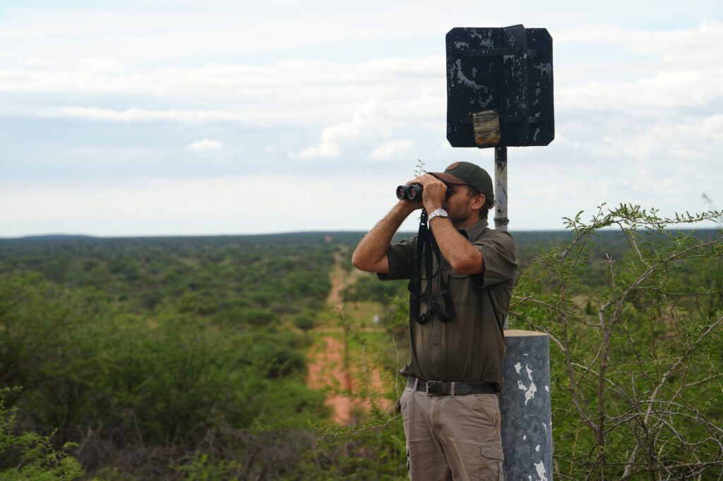 Mann mit Fernglas auf erhöhter Plattform blickt über die weite Savannenlandschaft Namibias – Wildbeobachtung auf einer Jagdfarm.