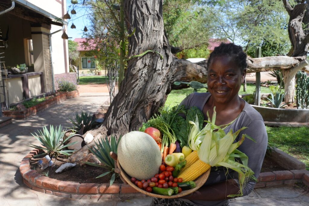 Lächelnde Frau auf einer namibischen Farm mit einem Korb frischem Gemüse aus eigenem Anbau – nachhaltige Landwirtschaft und Selbstversorgung in Afrika.